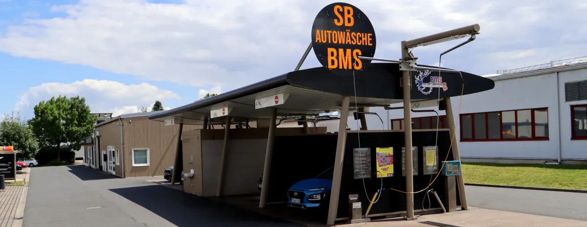 Überdachte Waschplätze der BMS SB-Waschanlage mit dem Schild „SB Autowäsche BMS“, im Waschplatz steht ein blaues Fahrzeug